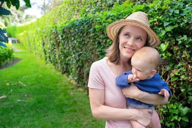 Dromerige moeder in hoed knuffelen schattige baby, glimlachend en wegkijken. schattige baby op moeder handen met vuist in de buurt van wang. familie zomertijd, tuin