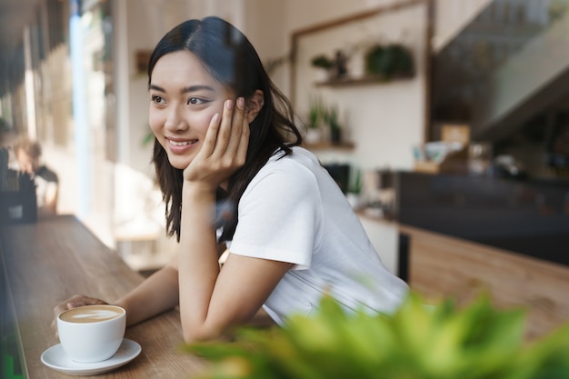 Dromerige mooie Aziatische meisje in casual wit t-shirt zit alleen stadscafé, koffie drinken.