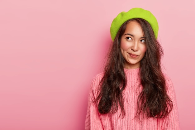 Dromerige mooie aziatische vrouw die diep in gedachten is, kijkt opzij en tuit lippen, draagt groene baret en gebreide trui