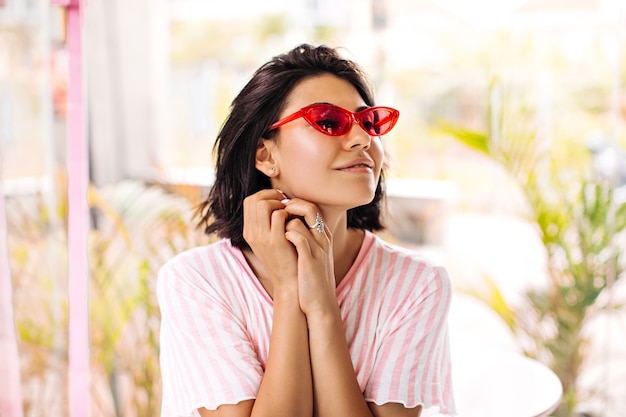 Dromerige stijlvolle dame met ring wegkijken op onscherpe achtergrond. Buiten schot van geïnspireerde brunette vrouw in roze zonnebril in de zomer.