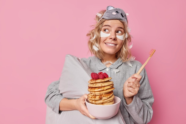 Dromerige tevreden jonge vrouw past schoonheidspleisters toe onder de ogen houdt tandenborstel en kom pannenkoeken poses met kussen draagt slaapmasker en pyjama geïsoleerd over roze achtergrond Hygiëne routines