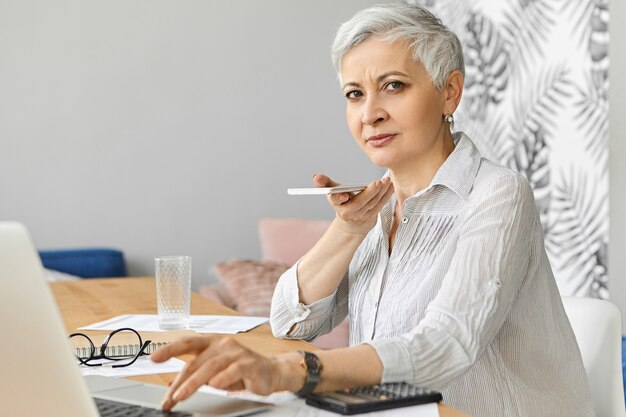 Drukke aantrekkelijke grijze haren blanke vrouw accountant over pensionering werkt als freelancer financiën beheren, zittend aan een bureau met draagbare computer, mobiele telefoon vasthouden, spraakbericht opnemen