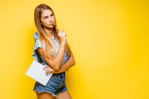 Drukke charmante aantrekkelijke vrouw die notities schrijft op notitieblok met trendy stijlvol blauw T-shirt geïsoleerd op gele muur