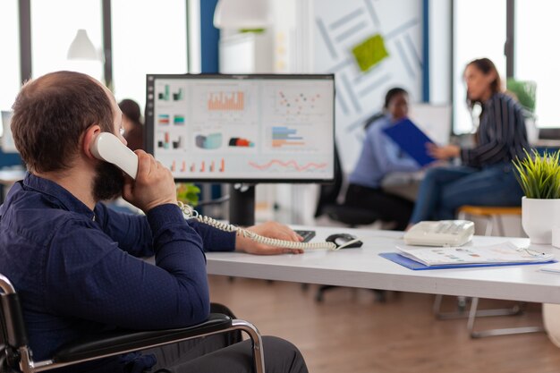 Drukke gehandicapte teamleider die de telefoon beantwoordt en praat met partner die op de computer typt, projectidee uitlegt en financiële gegevens controleert checking