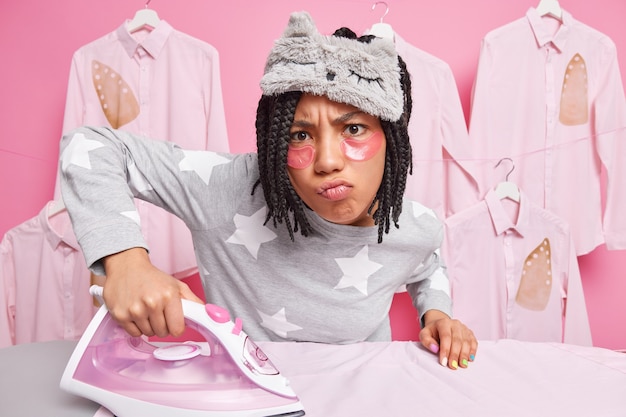 Gratis foto drukke ontevreden huisvrouw strijkt maakt ongelukkige grimas op camera gekleed in nachtkleding strijkijzers echtgenoten kleding poses tegen roze muur
