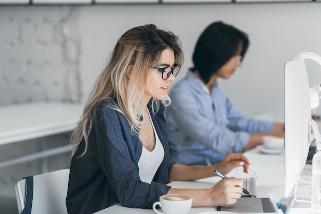 Drukke vrouwelijke freelancer met lang haar, werken met tablet en koffie drinken. Indoor portret van geconcentreerde Japanse student met behulp van computer in de klas.