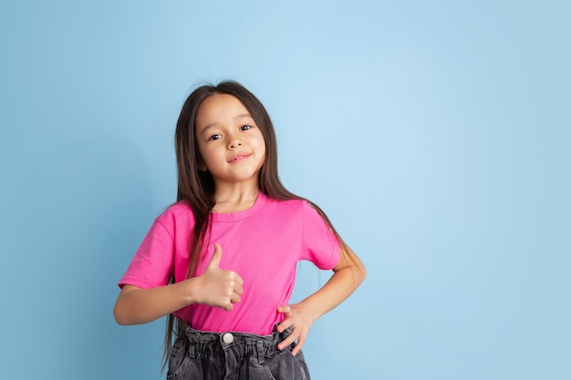 Duim omhoog, mooi gebaar. Het portret van het Kaukasische meisje op blauwe muur. Mooi vrouwelijk model in roze overhemd.