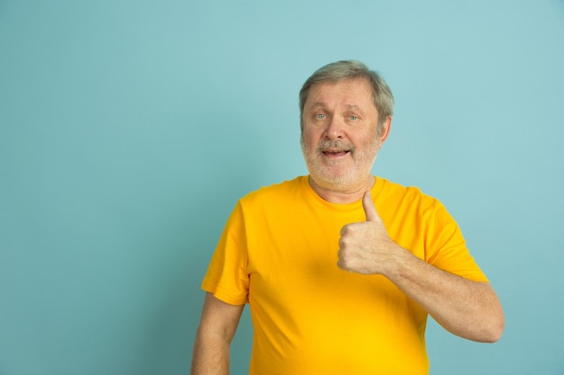 Duim opdagen, glimlachend. Blanke man portret geïsoleerd op blauwe studio achtergrond. Mooi mannelijk model in het gele overhemd stellen. Concept van menselijke emoties, gezichtsuitdrukking, verkoop, advertentie. Copyspace.