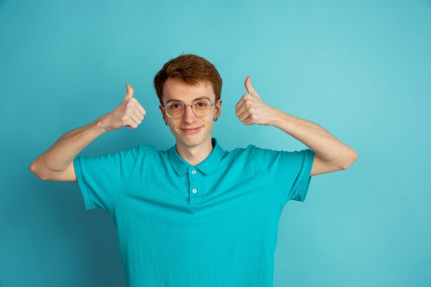 Duimen omhoog. Het moderne portret van de blanke jongeman geïsoleerd op een blauwe muur, zwart-wit. Mooi mannelijk model. Concept van menselijke emoties, gezichtsuitdrukking, verkoop, advertentie, trendy.