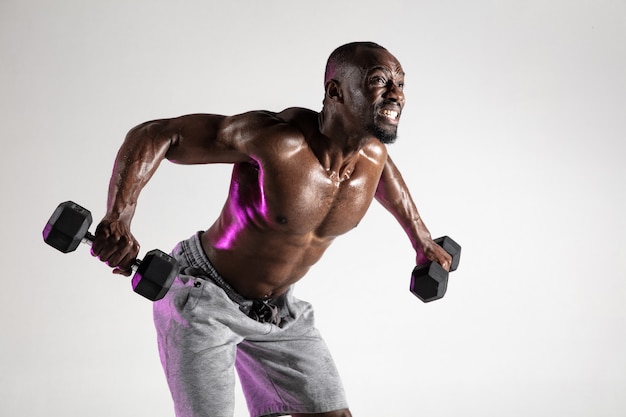 Dus de vleugels laten groeien. Jonge Afro-Amerikaanse bodybuilder training over grijze achtergrond. Gespierd enkel mannelijk model in sportkleding met gewichten. Concept van sport, bodybuilding, gezonde levensstijl.