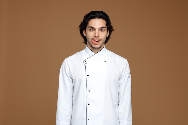 Gratis foto dwaze jonge mannelijke chef-kok in uniform kijkend naar camera met gekruiste ogen die tong tonen geïsoleerd op bruine achtergrond