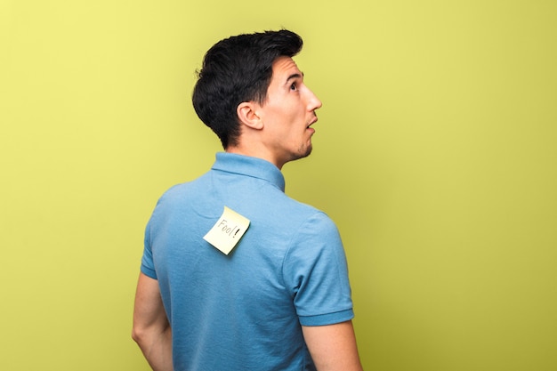 Dwaze uitziende man in blauw poloshirt met een gele kleverige nota met woord dwaas op zijn rug dom kijken Premium Foto