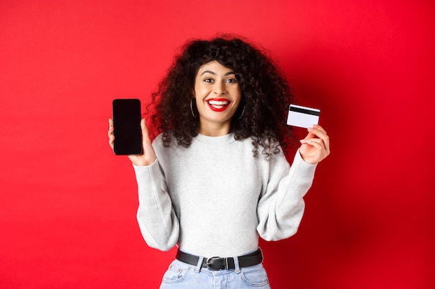 E-commerce en online winkelconcept. Vrolijke vrouw die lacht, met plastic creditcard en leeg smartphonescherm, staande op rode achtergrond.