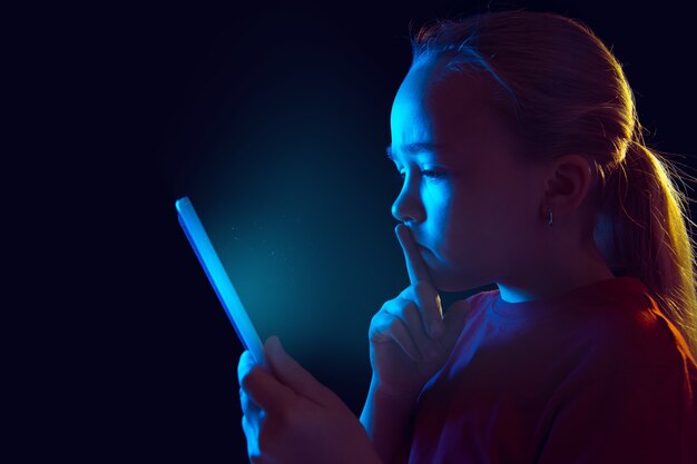 Echt. Het portret van het Kaukasische meisje dat op donkere muur in neonlicht wordt geïsoleerd. Mooi vrouwelijk model dat tablet gebruikt. Concept van menselijke emoties, gezichtsuitdrukking, verkoop, advertentie, moderne technologie, gadgets.