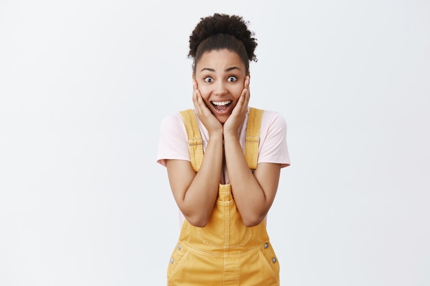 Echt niet, beroemdheid in mijn buurt. portret van gecharmeerd verbaasd gelukkig donker gekleurd vrouwelijk model in gele overall, kin aanraken met handpalmen en vreugdevol glimlachen, schreeuwen van verbazing