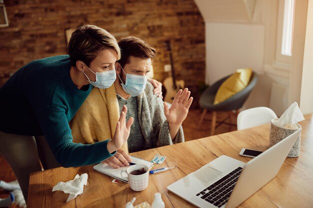 Echtpaar met beschermende gezichtsmaskers die tijdens videogesprek over laptop thuis zwaaien