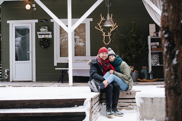 Echtpaar van middelbare leeftijd zittend op een besneeuwd terras stapt voor het huis en poseert voor een foto. beiden dragen hoeden, jassen en sjaals. hij houdt haar enkels op zijn knieën.