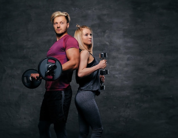 Een aantrekkelijk fitnesspaar van sportieve mannen houdt een halter vast en een slanke blonde vrouw houdt halters vast over een grijze achtergrond