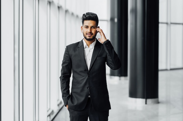 Een aantrekkelijke Indiase zakenman met een baard in een modern kantoorcentrum, gekleed in een zwart pak, houdt zijn hand bij een draadloze oortelefoon