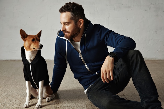 Een aantrekkelijke jonge man en zijn bassenji-hond in bijpassende hoodies zitten op de grond naast een witte muur