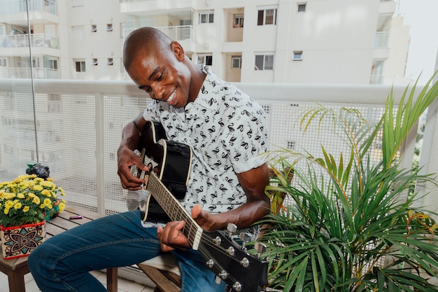 Een Afrikaanse lachende jonge man zittend op een stoel in het balkon genieten van gitaar spelen
