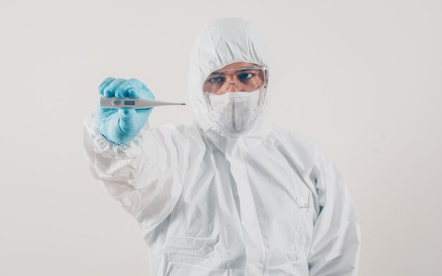 Een arts die en thermometer op lichte achtergrond in masker, medische handschoenen en beschermend kostuum bevindt zich houdt. coronavirus