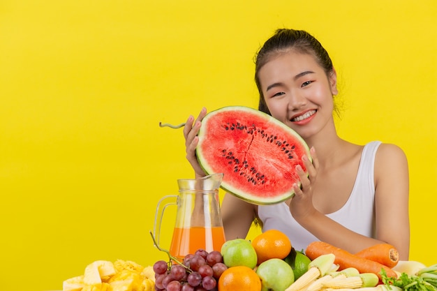 Een Aziatische vrouw die een wit mouwloos onderhemd draagt. Beide handen houden watermeloenen vast en de tafel staat vol met verschillende soorten fruit.