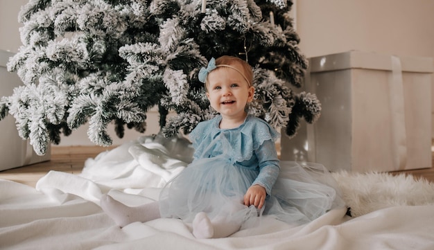 Een babymeisje in een blauwe feestjurk zit op een witte plaid bij de kerstboom en kijkt in de camera