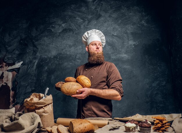 Een bebaarde bakker in een uniform met vers brood, staande naast een tafel, versierd met heerlijke broden, baguettes en muffins in een donkere studio