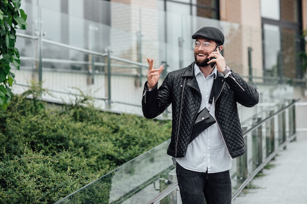 Een bebaarde, glimlachende, stijlvolle manager die telefoon spreekt in de straten van de stad in de buurt van een modern kantoor