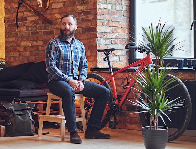 Een bebaarde hipster man gekleed in een fleece shirt zit op een houten stoel, bij het raam in een kamer met loft interieur