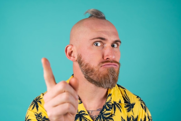 Een bebaarde man in een overhemd met een print van palmbomen op een turquoise muur kijkt verwijtend naar voren