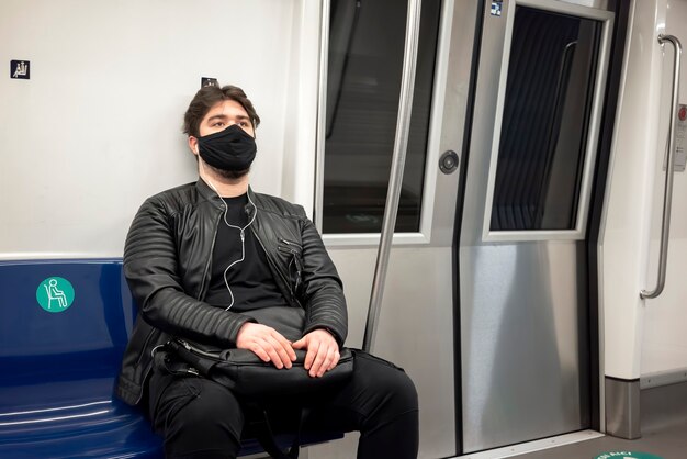 Een blanke man met baard en koptelefoon in zwart medisch masker zittend op een stoel in de metro