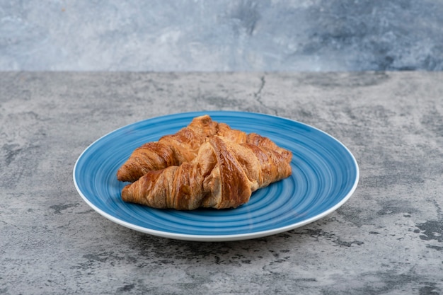 Een blauw bord van twee eenvoudige verse croissants op een stenen tafel.