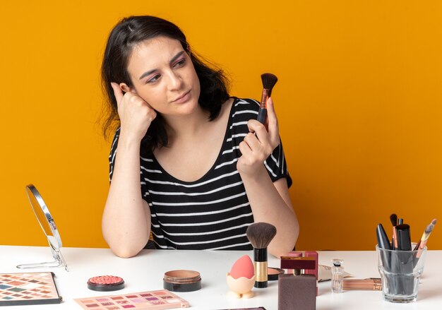 Een blij jong mooi meisje zit aan tafel met make-uptools die een poederborstel vasthouden en bekijken die op een oranje muur is geïsoleerd