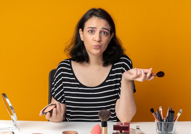 Een blij jong mooi meisje zit aan tafel met make-uptools met een borstel met poederblush geïsoleerd op een oranje muur