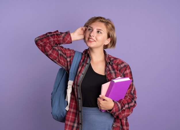 Een blij jong slavisch studentenmeisje dat een rugzak draagt, legt de hand op het hoofd, houdt een boek vast en kijkt naar de zijkant