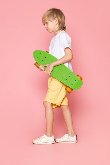 Een blonde blonde jongen van het vooraanzicht in witte t-shirt en gele jeans die groen skateboard op de roze ruimte houden