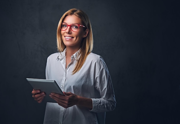 Een blonde vrouw gekleed in een wit overhemd en een bril houdt tablet-pc op een grijze achtergrond.