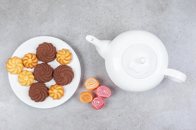Een bord koekjes naast een witte theepot en een klein bundeltje marmelade op een marmeren oppervlak.