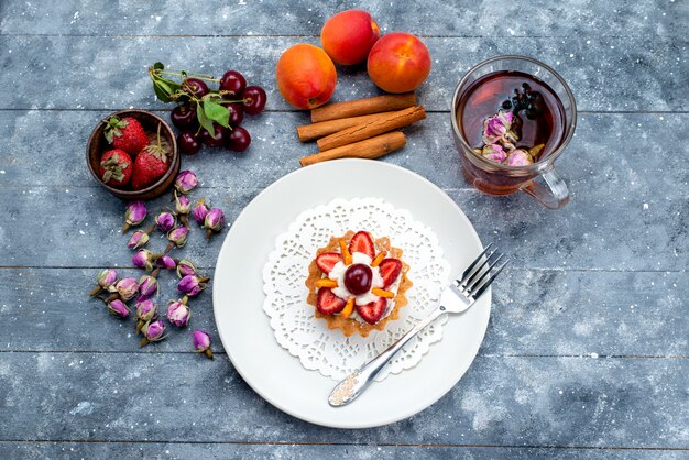 Een bovenaanzicht heerlijke kleine cake met room en vers gesneden fruit samen met kaneelthee op het grijsblauwe bureau fruitcake biscuit thee