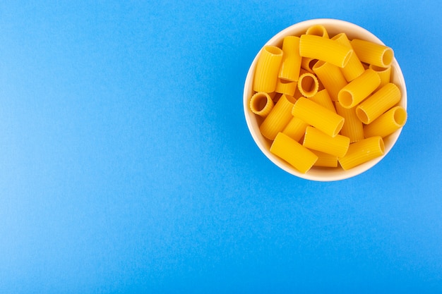 Een bovenaanzicht Italiaanse droge pasta vormde kleine gele rauwe pasta binnen crème gekleurde ronde kom geïsoleerd op de blauwe