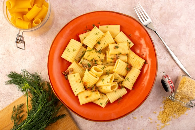 Een bovenaanzicht Italiaanse pasta gekookt lekker gezouten binnen ronde oranje plaat met greens en rauwe pasta op roze bureau