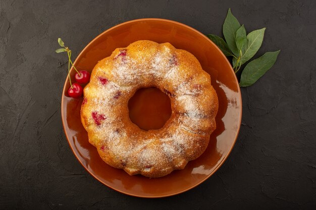 Een bovenaanzicht kersencake ronde gevormd binnen bruine plaat op het donkere bureau cake koekje suiker zoet