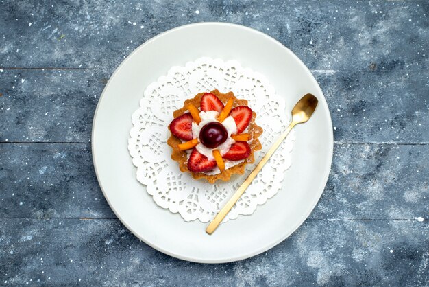 Een bovenaanzicht kleine heerlijke cake met room en fruit in een witte plaat op het grijsblauwe fruitcake van het bureau