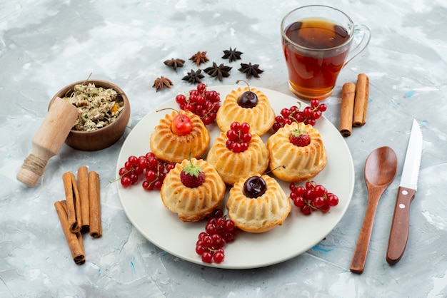 Een bovenaanzicht lekkere kleine cakes in een witte plaat met kersen en aardbeien samen met kaneel en thee op de lichttafel cake fruit bes