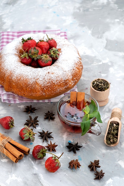 Een bovenaanzicht ronde cake met suikerpoeder rode aardbeien kaneelthee op de witte bessencake van het bureau