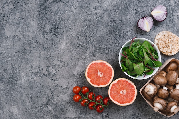 Een bovenaanzicht van champignons; grapefruit slice; ui; spinazie; kerstomaatjes en gepofte rijst cake op grijze gestructureerde achtergrond