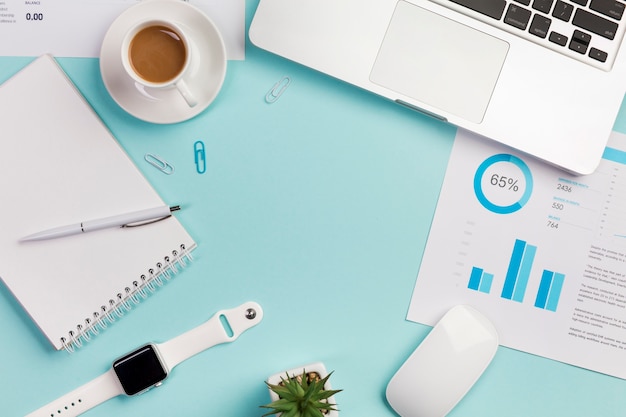 Een bovenaanzicht van een bureau met briefpapier, laptop, muis en slimme horloge op blauwe achtergrond