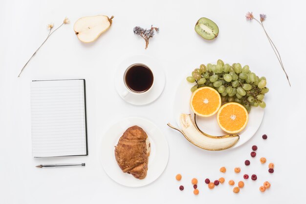 Een bovenaanzicht van een notebook; pen; croissant; vruchten; koffie en gedroogde bloemen op witte achtergrond